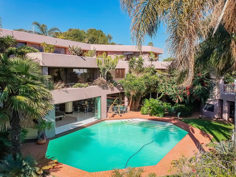 An elevated view of a multi-level residential property featuring a swimming pool, brick patio area, and mature landscaped garden.