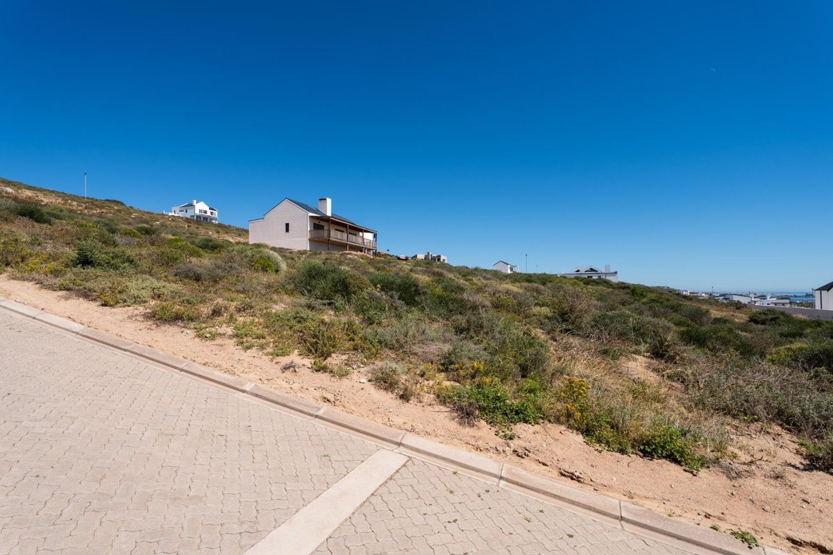 A view of a vacant residential plot featuring sloped terrain, adjacent to a paved street with concrete curbing in a coastal development.