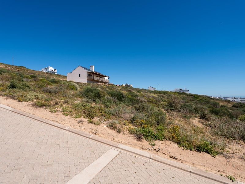 A view of a vacant residential plot featuring sloped terrain, adjacent to a paved street with concrete curbing in a coastal development.