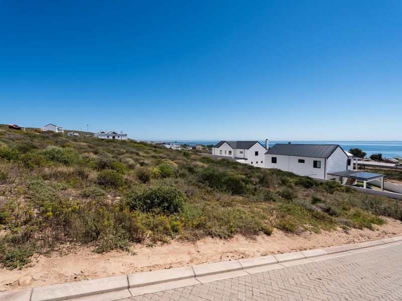 A scenic view of a vacant residential plot featuring natural coastal vegetation, paved road access, and distant ocean views.