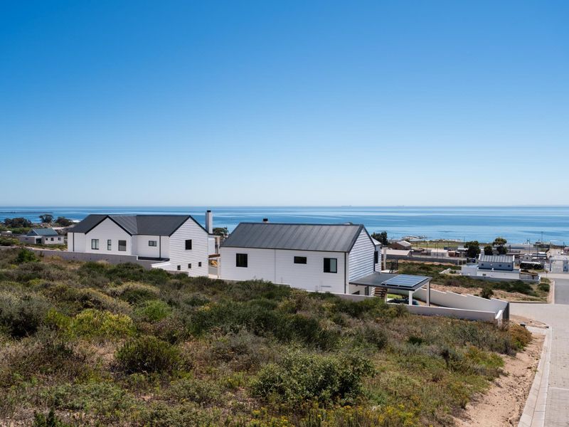 Modern coastal residential property featuring white siding, dark metal roofing, solar panels, and a paved driveway with expansive ocean views.