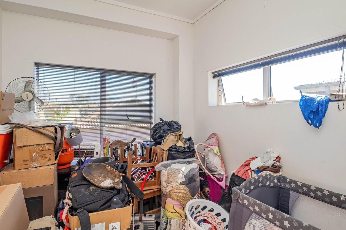 A bedroom space featuring aluminium window frames and venetian blinds, currently used for storage with visible wall plaster cracking.