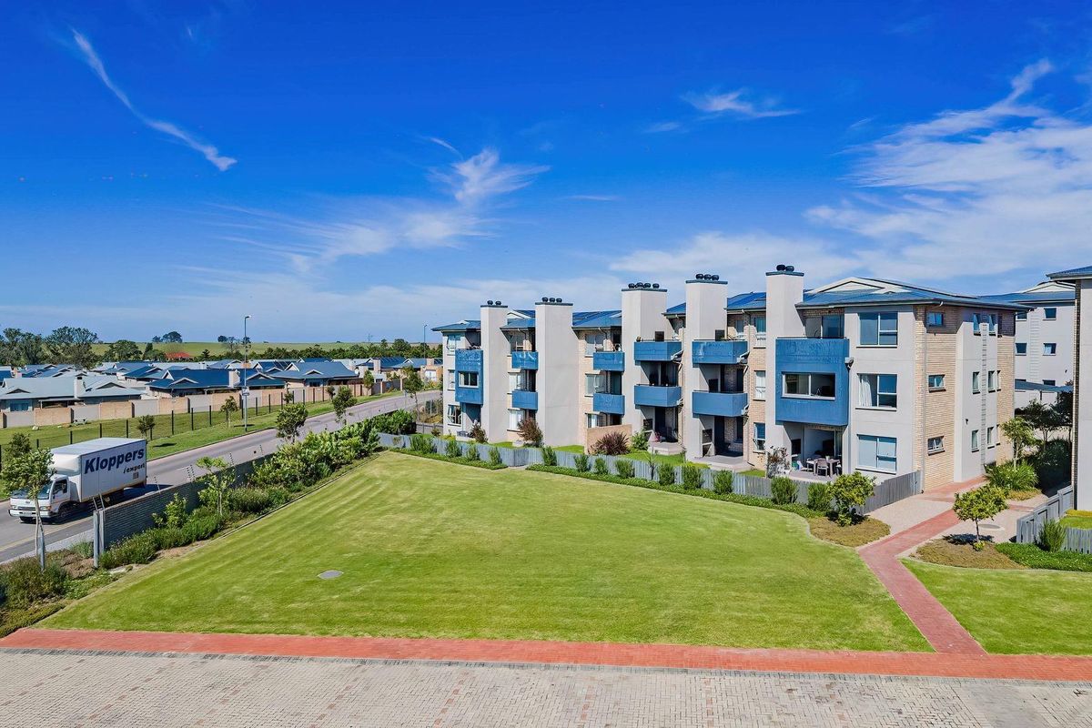 An aerial view of a modern multi-story apartment complex featuring landscaped lawns, paved pathways, and private balconies under a clear blue sky.