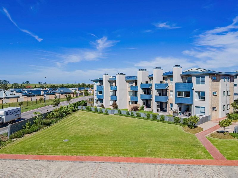 An aerial view of a modern multi-story apartment complex featuring landscaped lawns, paved pathways, and private balconies under a clear blue sky.