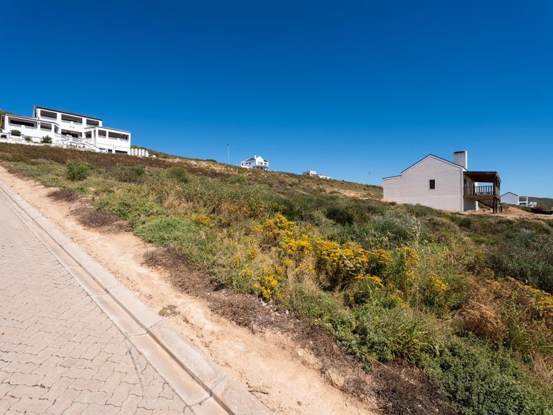 A view of a sloped residential vacant land plot adjacent to a paved road in a coastal neighborhood.