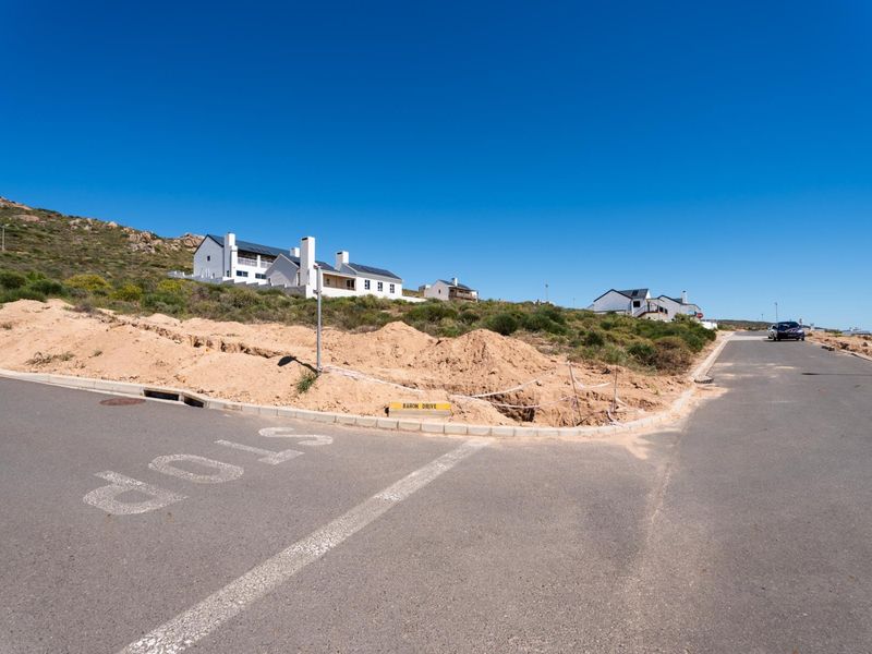 Street view of a residential corner plot featuring asphalt road access, concrete curbing, and an excavated soil mound on the verge.