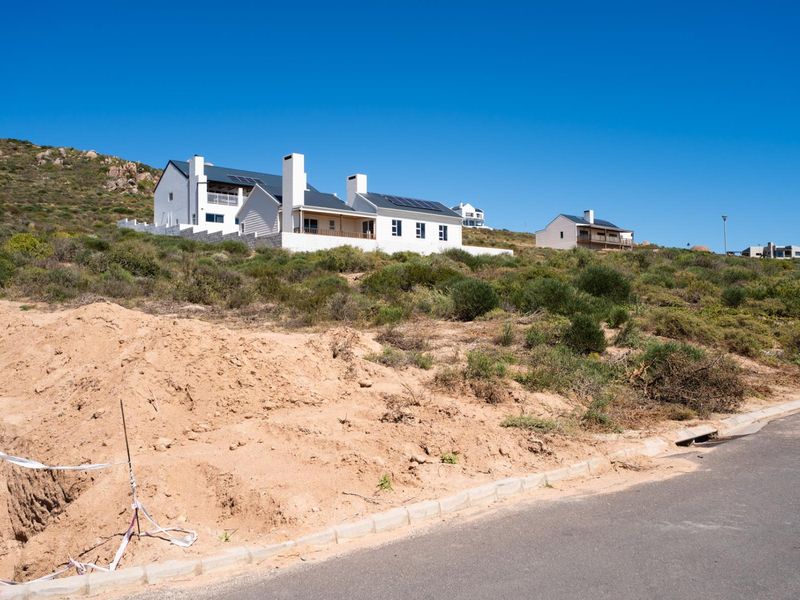 A view of a vacant residential plot featuring natural vegetation and an excavated soil mound, with established houses visible in the background.