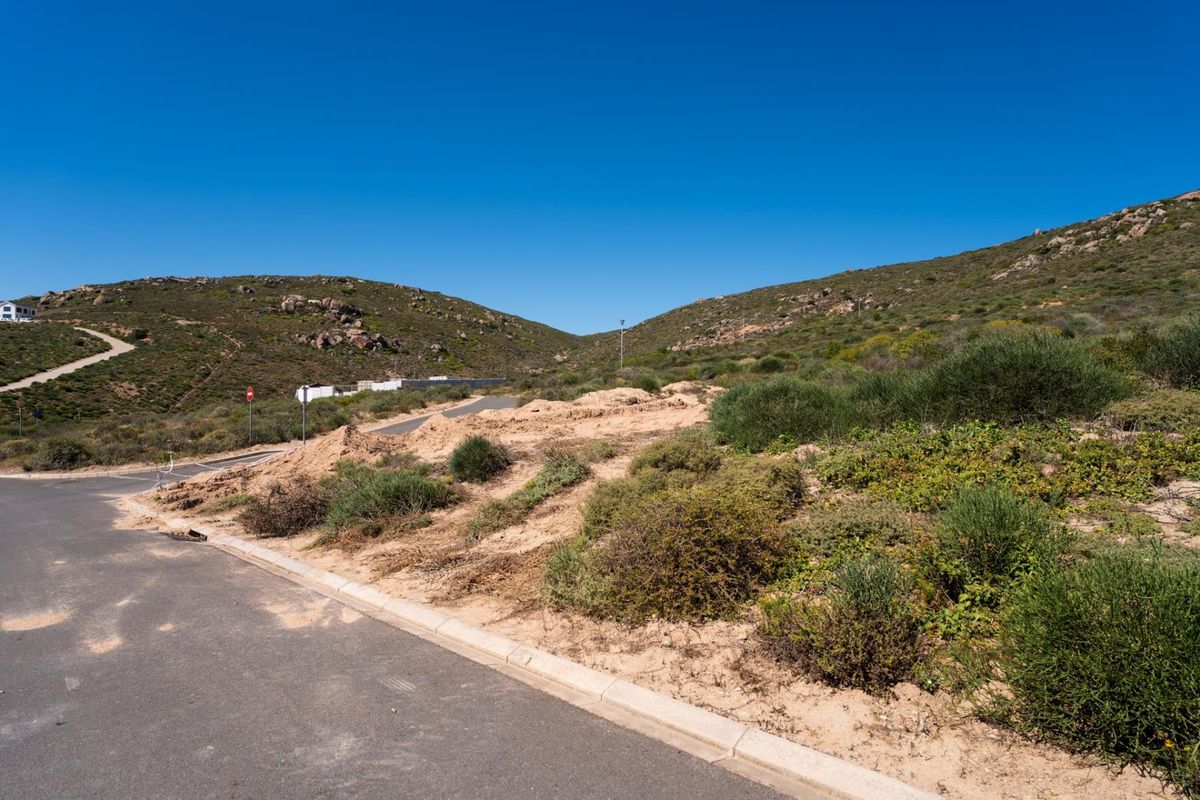 A vacant residential plot featuring sloped natural terrain, accessible via a paved road with concrete curbing in a quiet hillside setting.