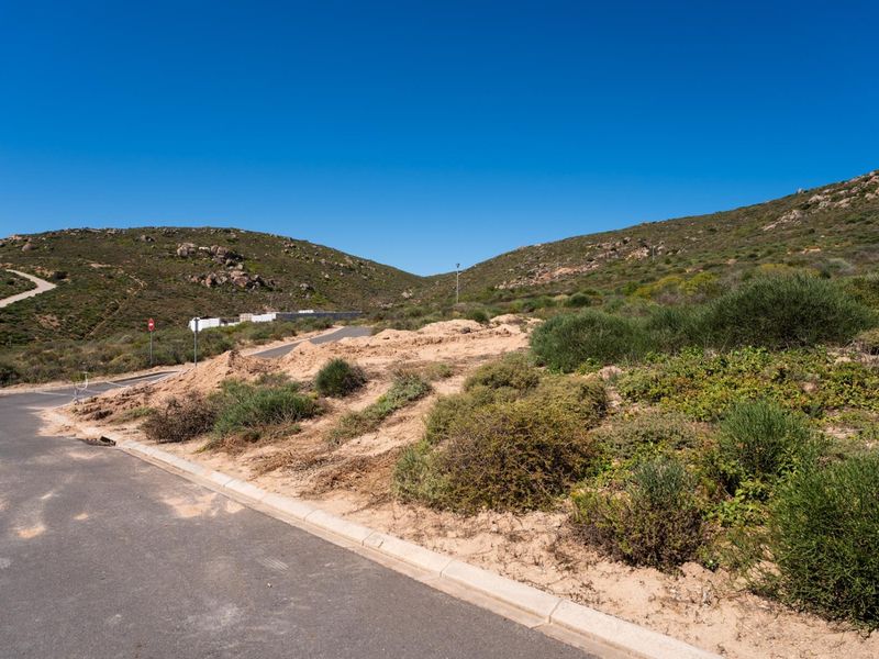 A vacant residential plot featuring sloped natural terrain, accessible via a paved road with concrete curbing in a quiet hillside setting.