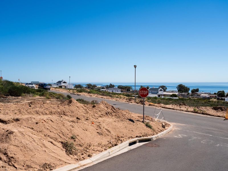 A street view of the vacant residential land showing the paved road, curbing, and surrounding coastal landscape.