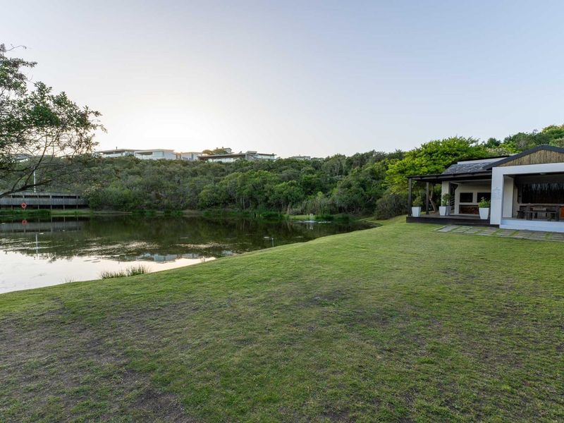 A scenic rear exterior view featuring a well-maintained lawn, a natural pond, and a covered patio area with a built-in braai.
