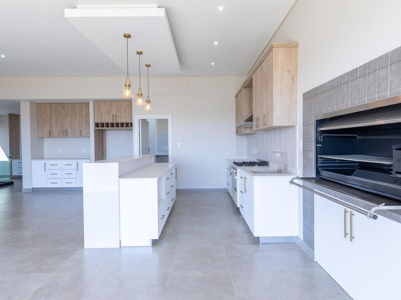 Modern open-plan kitchen and entertainment area featuring a built-in braai, central island, gas hob, and sleek cabinetry with neutral tiled flooring.