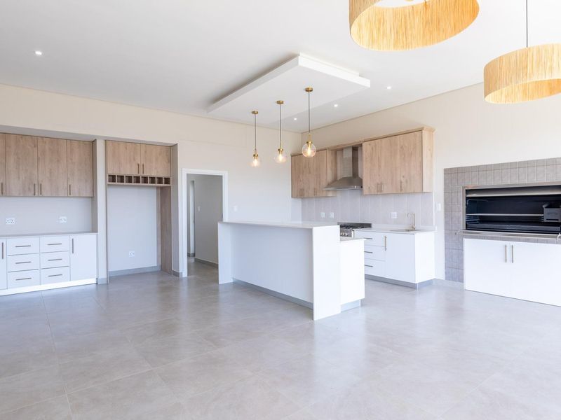 Modern open-plan kitchen and living area featuring sleek cabinetry, a central island, built-in braai, and neutral tiled flooring throughout.