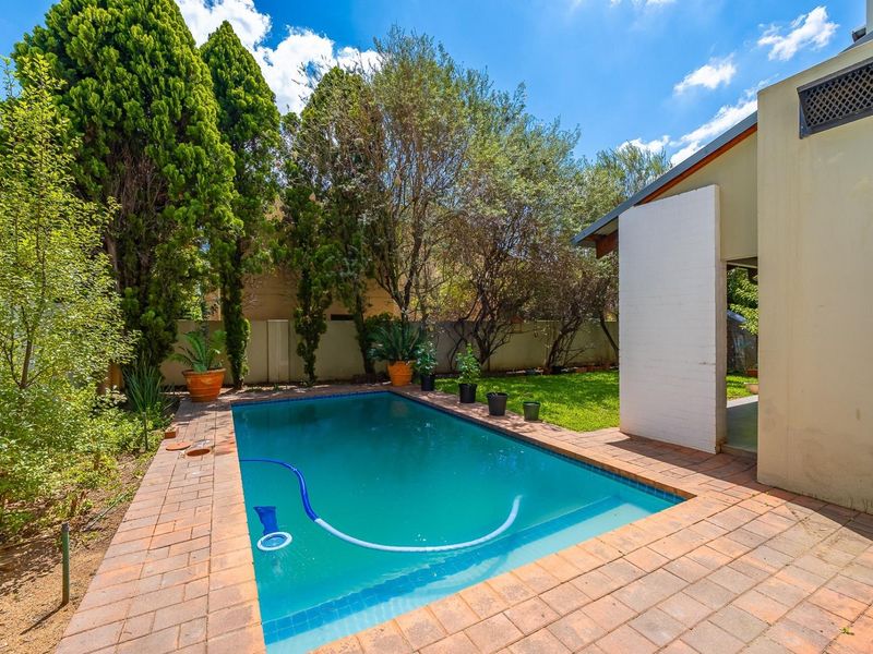 A private backyard featuring a swimming pool with brick paving surround, established trees, and a boundary wall.