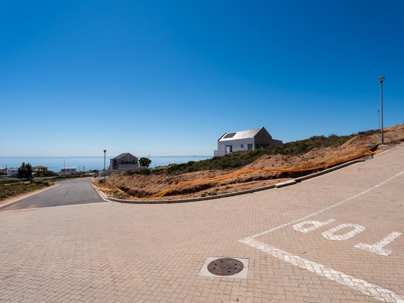 A view of a residential street with paved surfaces, infrastructure, and a vacant plot overlooking the ocean in a coastal development.