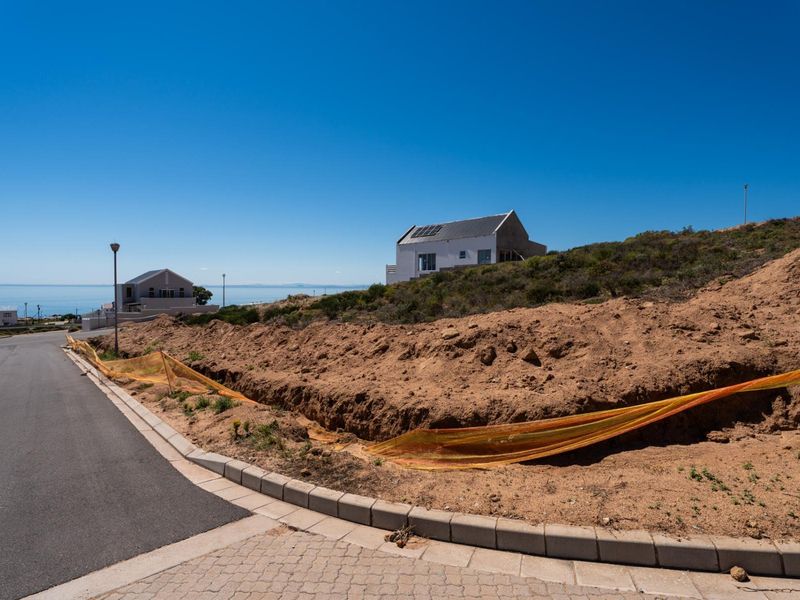 A view of a vacant residential plot featuring sloped terrain, adjacent to a paved street with concrete curbing in a coastal neighborhood.