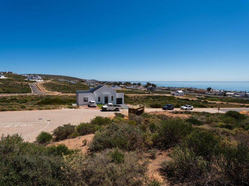 An elevated aerial view of a residential coastal area featuring natural vegetation, paved roads, and distant ocean views.