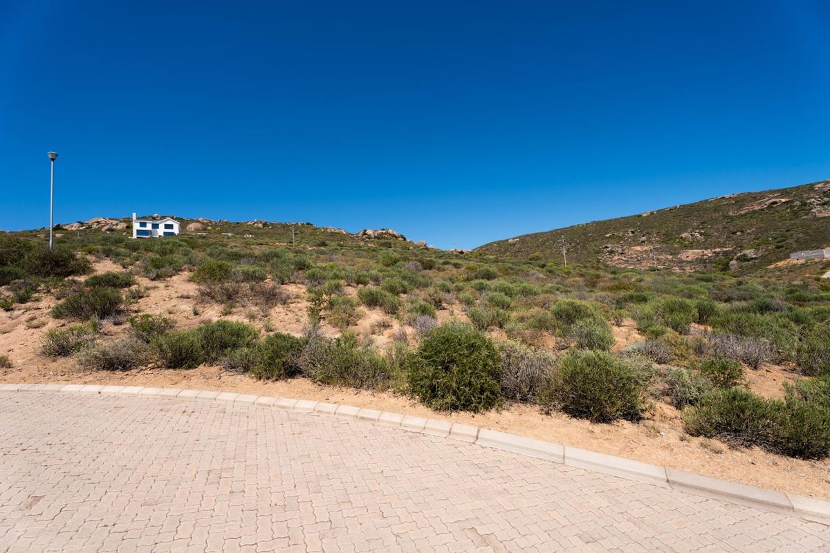 A view of a vacant residential plot featuring a paved driveway, kerbing, and natural hillside vegetation under a clear blue sky.