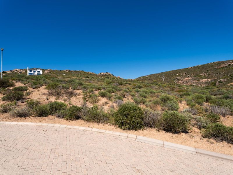 A view of a vacant residential plot featuring a paved driveway, kerbing, and natural hillside vegetation under a clear blue sky.