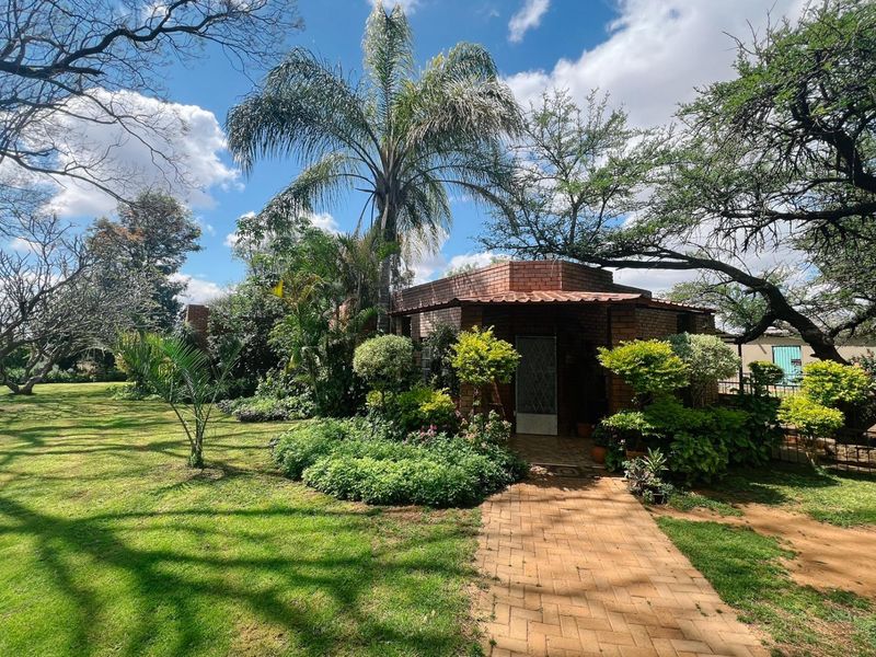 A charming brick-faced farm cottage entrance featuring a paved walkway, established garden landscaping, and mature trees under a bright blue sky.