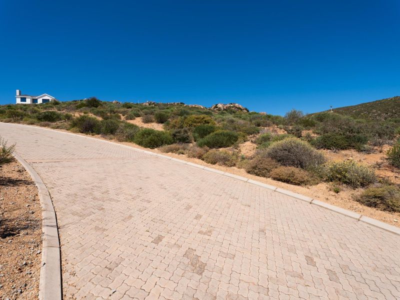 A paved residential driveway leading up a gentle slope, surrounded by natural coastal vegetation and clear blue skies.