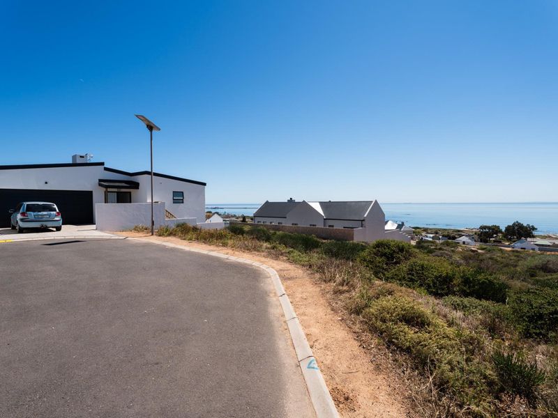 A street-level view of the residential property showing the paved road, curb, and solar street lighting with a coastal landscape in the background.
