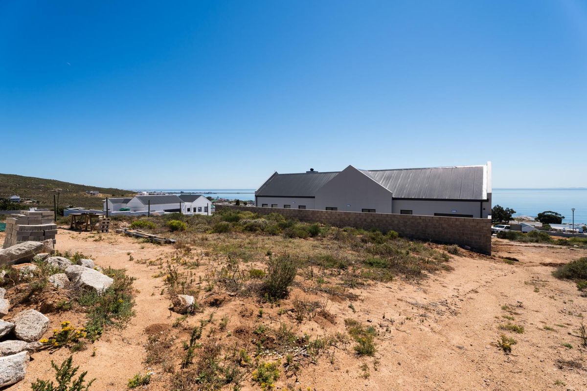 A view of a vacant residential plot featuring natural vegetation and a boundary wall, with neighboring properties and coastal views in the background.