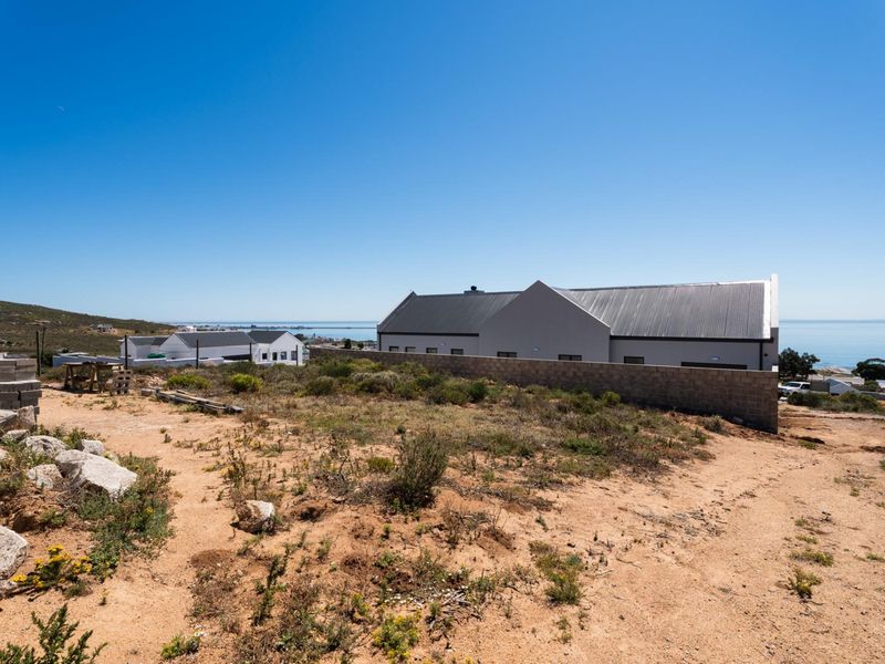 A view of a vacant residential plot featuring natural vegetation and a boundary wall, with neighboring properties and coastal views in the background.