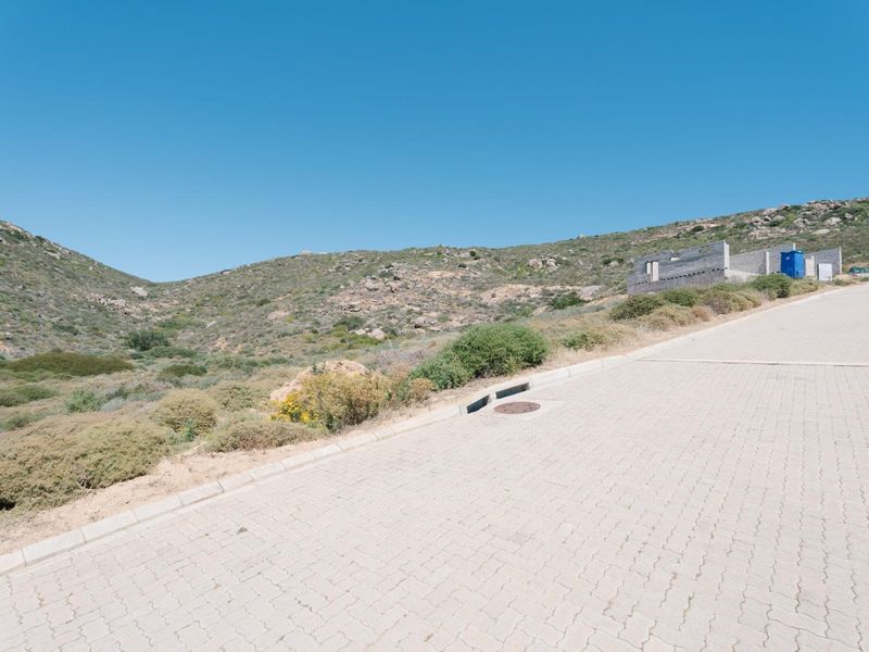 A view of a paved residential street with kerbing and stormwater infrastructure, set against a natural hillside landscape.