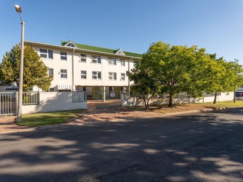 Front exterior view of a multi-story apartment complex featuring a green metal roof, secure perimeter fencing, and a paved entrance area.