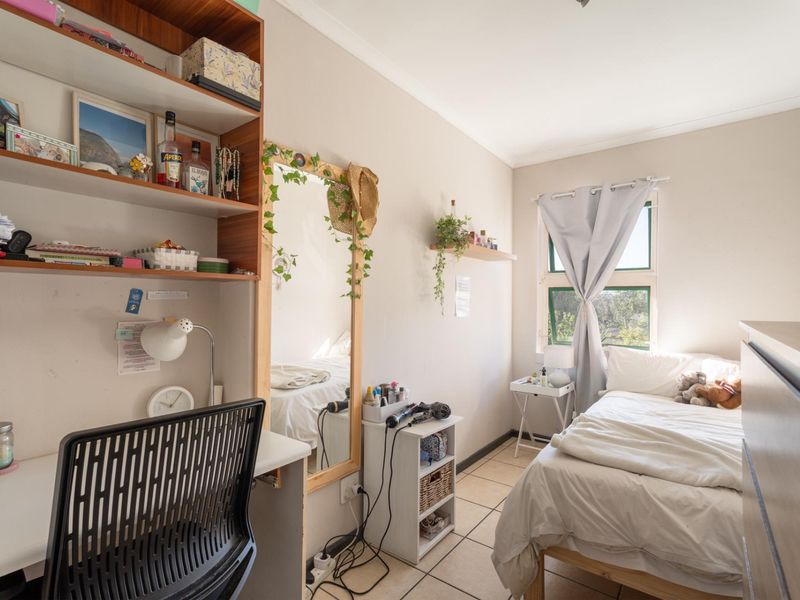 A functional bedroom featuring tiled flooring, a built-in desk with shelving, and a window providing natural light.
