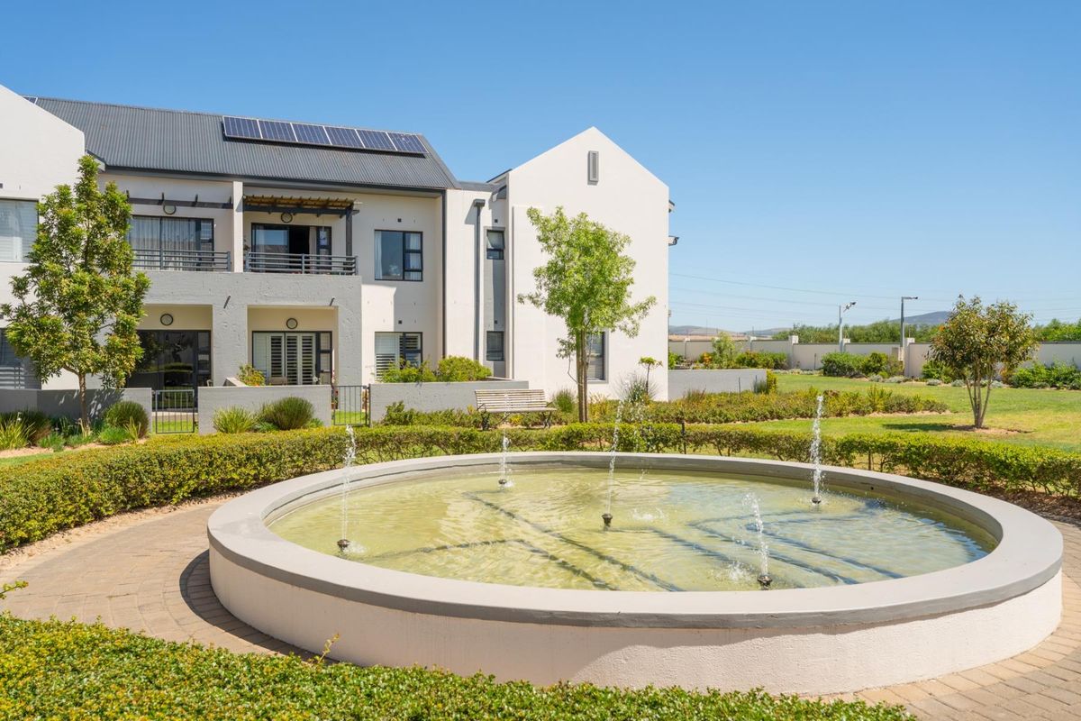 A well-maintained apartment complex exterior featuring a central water fountain, landscaped gardens, and private balconies with solar panels installed on the roof.