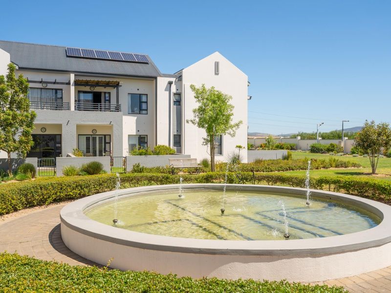 A well-maintained apartment complex exterior featuring a central water fountain, landscaped gardens, and private balconies with solar panels installed on the roof.