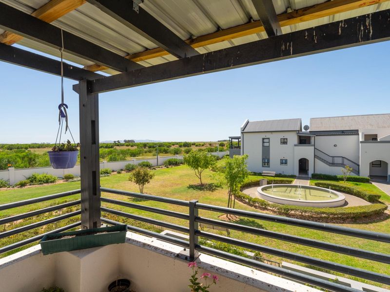 A covered balcony featuring a timber pergola and metal railing, overlooking a landscaped garden with a central water fountain and apartment complex views.