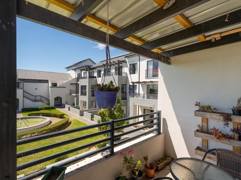 A covered balcony featuring a timber pergola and metal balustrade, overlooking the apartment complex courtyard and garden area.