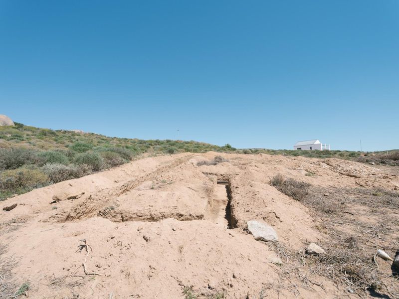 A view of a vacant residential plot featuring an excavated trench in the sandy terrain under a clear blue sky.