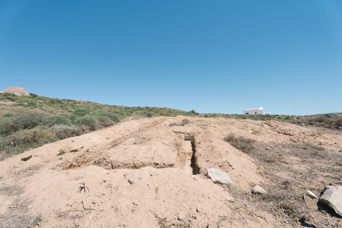 A view of a vacant residential plot featuring an excavated trench in the sandy terrain under a clear blue sky.