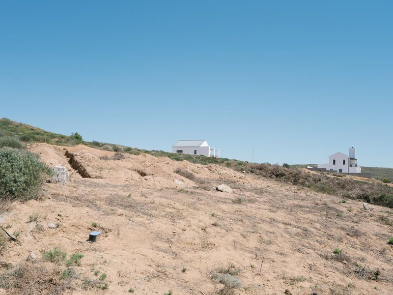 A view of a sloped, undeveloped residential plot with natural vegetation and surrounding neighborhood homes in the distance.