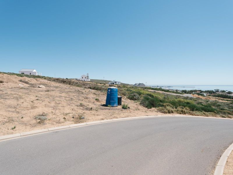 A view of a vacant residential plot from the adjacent paved street, featuring natural coastal terrain and distant ocean views.