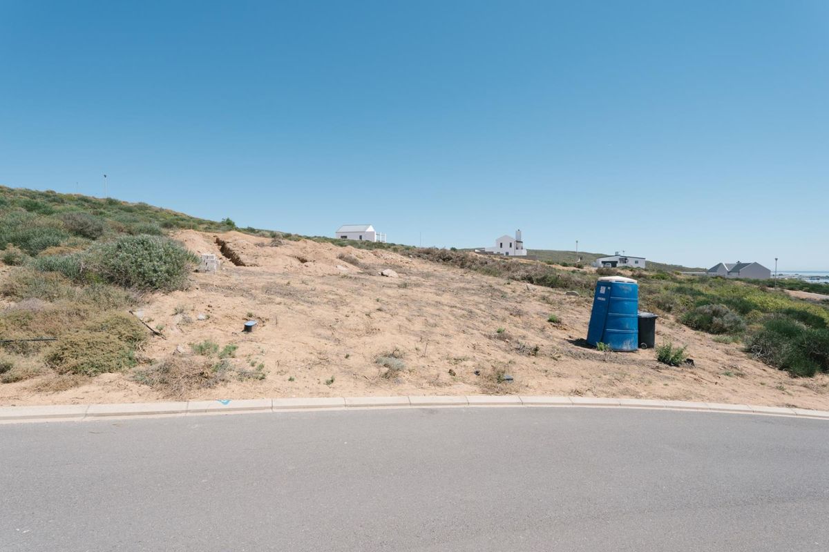 A vacant residential plot situated on a paved street, featuring natural coastal vegetation and a gentle slope, ready for development.