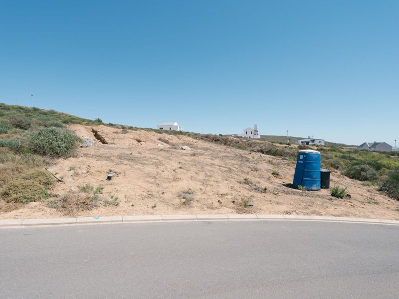 A vacant residential plot situated on a paved street, featuring natural coastal vegetation and a gentle slope, ready for development.