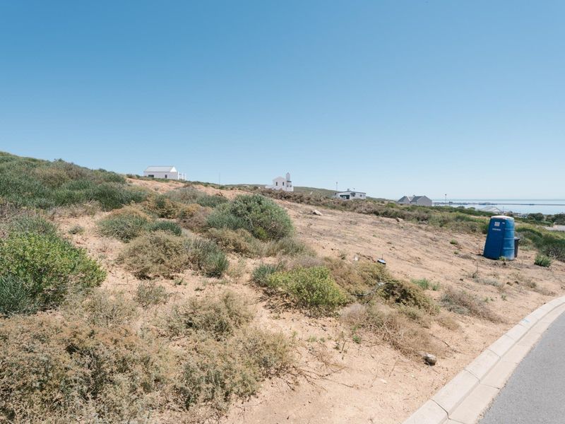 A vacant residential plot featuring natural coastal vegetation, sloping terrain, and direct access to a paved street in a quiet neighborhood.