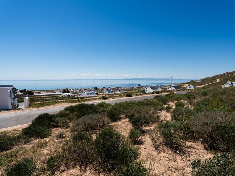 Elevated view of a vacant residential plot overlooking the coastline and surrounding neighborhood, featuring natural vegetation and paved road access.