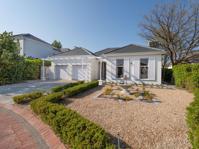 A well-maintained single-story home featuring a double garage, pergola, shuttered windows, and a landscaped front garden with a central stone fountain.