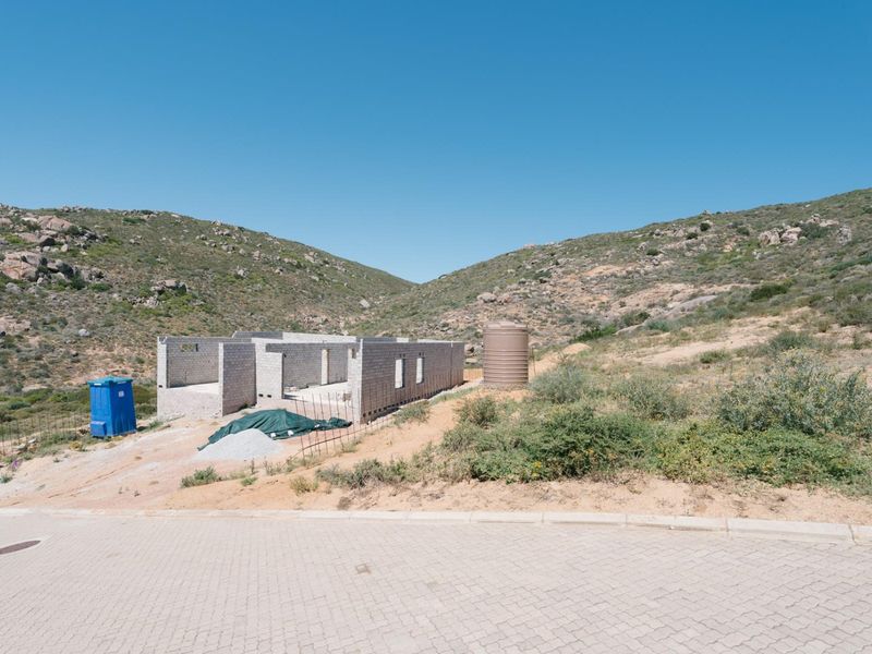 A residential plot featuring a partially constructed concrete block building structure and a water storage tank, set against a natural hillside landscape.