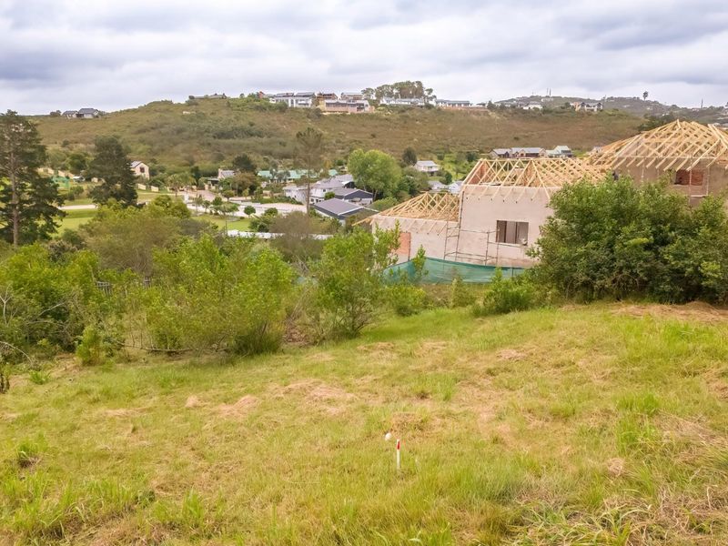 An elevated view of a vacant residential plot featuring natural grass terrain with a neighboring property currently under construction.