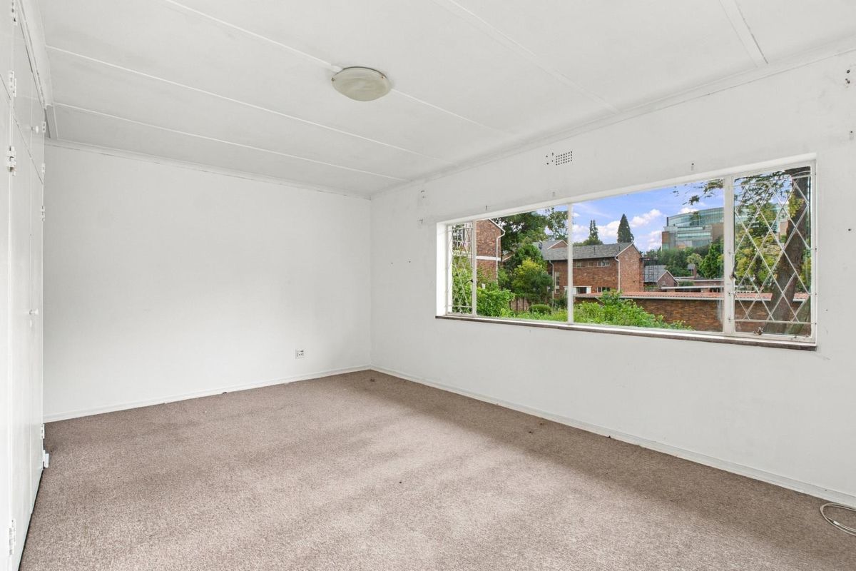 An empty bedroom featuring built-in cupboards, carpeted flooring, and a large window providing natural light and a view of the surrounding neighborhood.