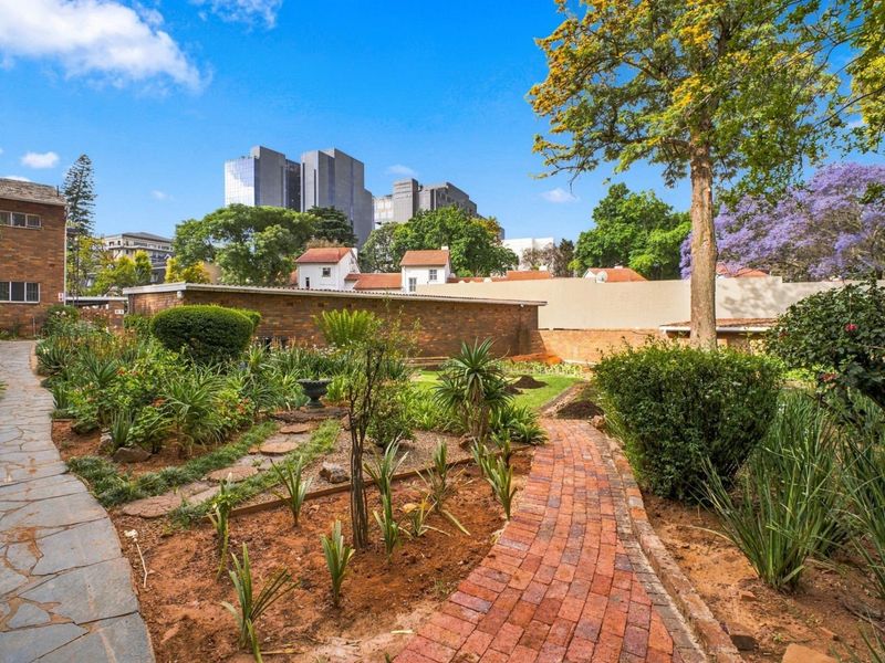 A well-maintained communal garden area featuring stone and brick pathways, established garden beds, and mature trees with urban views.