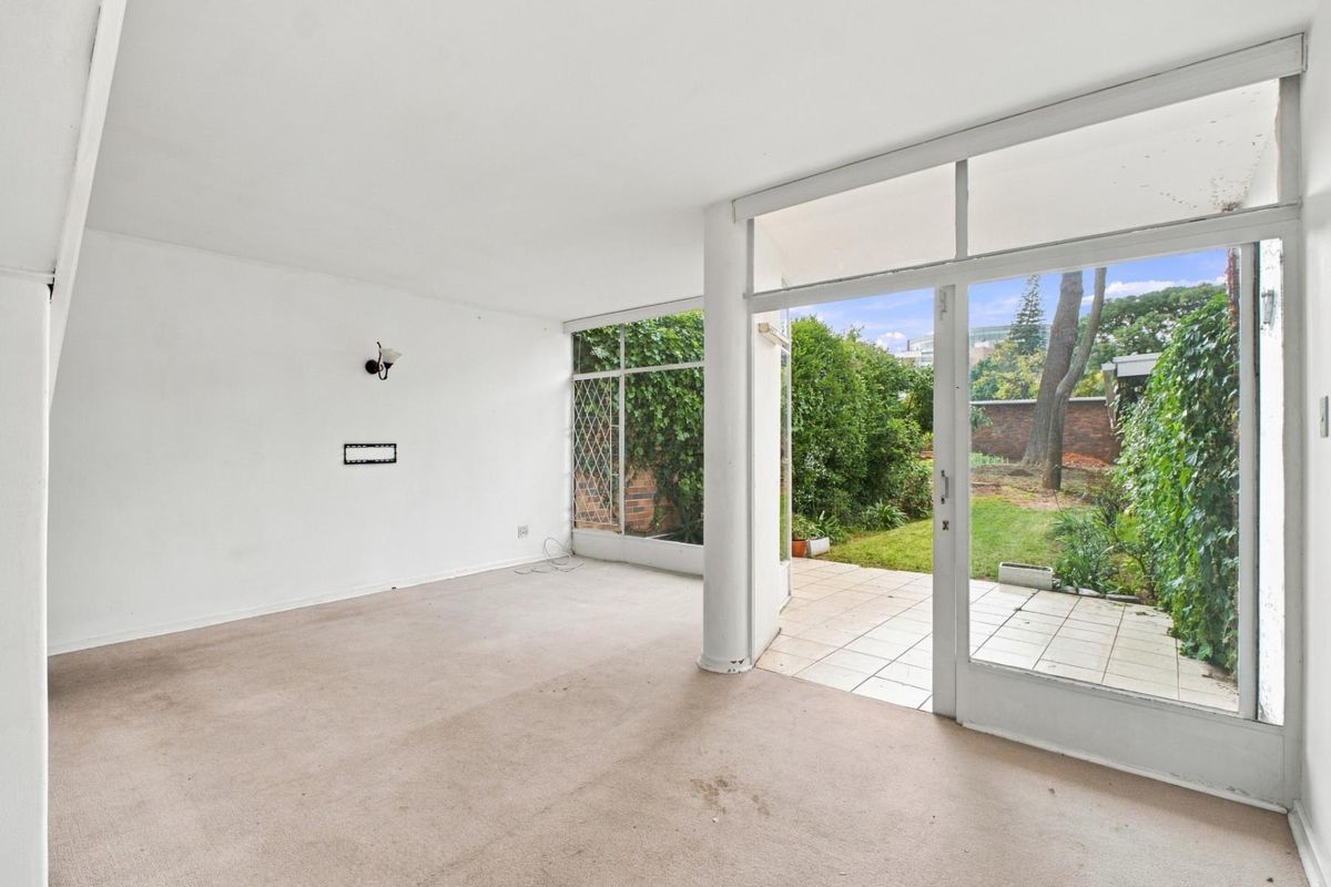An empty, carpeted living area featuring large glass sliding doors that open onto a private tiled patio and garden space.