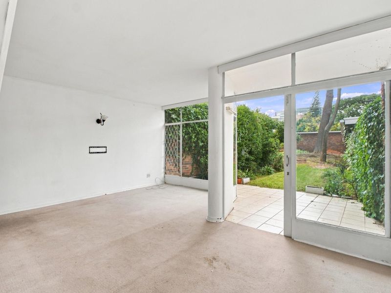 An empty, carpeted living area featuring large glass sliding doors that open onto a private tiled patio and garden space.